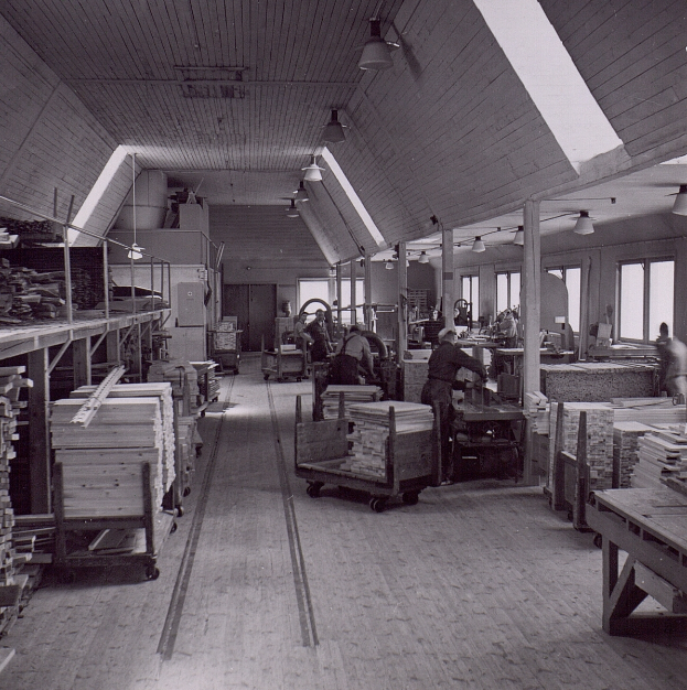 Ein Schwarz-Weiß-Foto eines Fabrikinneren mit Arbeitern, Tischen, mit Holz gefüllten Wagen und Deckenlampen sowie großen Fenstern an den Wänden.
