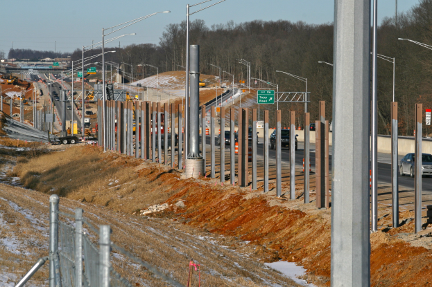 Baustelle mit Fahrzeugen, Polen, Lampen, Texttafeln, einem Zaun, schneebedecktem Gras, Bäumen und einem Himmel im Hintergrund.