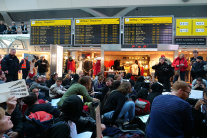 Eine große Gruppe von Menschen sitzt und steht in einem Flughafen während einer Demonstration, mit einigen haltenden Taschen und Papieren, während Hintergrundtafeln Text anzeigen, Puppen Kleider tragen und Deckenlichter die Szene beleuchten.