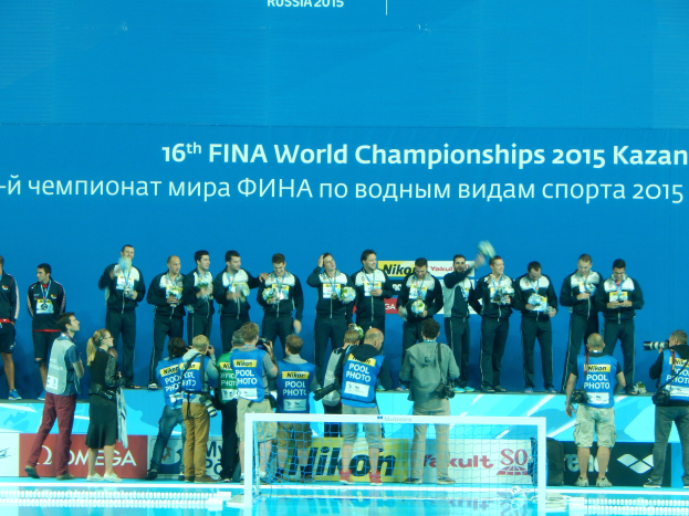 Eine Gruppe von Menschen, die auf einem Swimmingpool stehen, einige halten Kameras, mit einem Netz im Vordergrund und einem Banner im Hintergrund mit der Aufschrift "FINA World Championships 2015 Kazan".