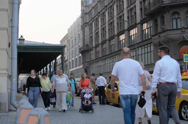 Eine Gruppe von Menschen, die eine Straße mit hohen Gebäuden entlanggehen, einige tragen Taschen und einer schiebt einen Kinderwagen, mit Fahrzeugen, einem Schild, Straßenlaternen, einer Metallhütte und einem bewölkten Himmel.