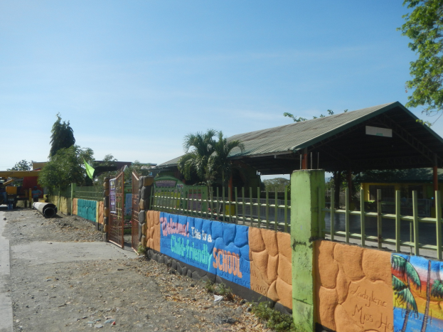 Schulgebäude mit Wandgemälde, umgeben von einem Zaun, einer Hütte, Bäumen, Fahrzeugen, Strommasten und einem bewölkten Himmel.
