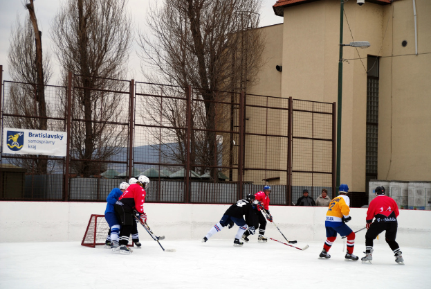 Personen beim Eisschnelllauf auf einer Eisfläche mit Gebäuden, Bäumen, einer Straßenlaterne, einem Namensschild und Zäunen im Hintergrund unter einem Himmel.