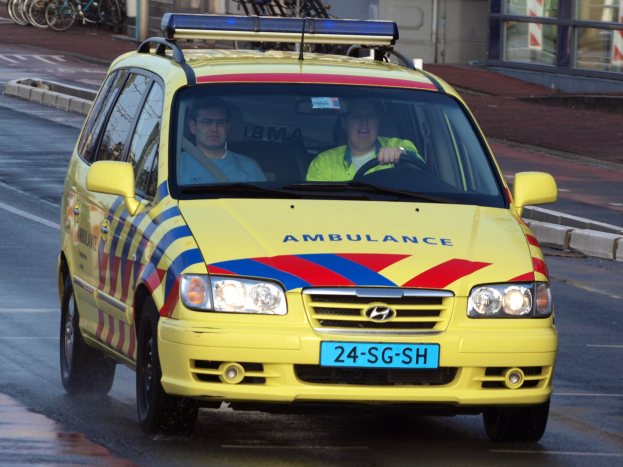 Ein gelber Krankenwagen mit zwei sichtbaren Insassen fährt auf einer Straße, mit parkenden Fahrrädern auf dem linken Gehweg und einem Glasfenster-Gebäude im Hintergrund.