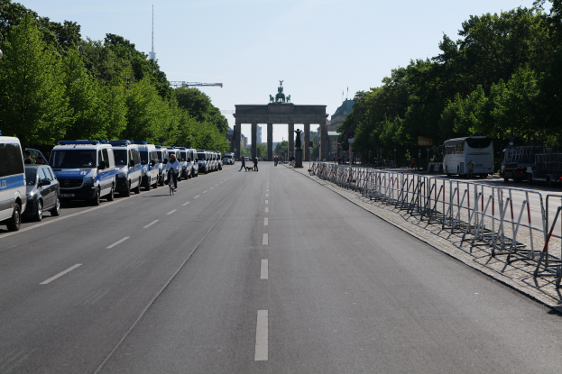 Eine Reihe von Polizeiwagen auf einer Straße vor dem Brandenburger Tor in Berlin, Deutschland geparkt, mit Menschen auf Fahrrädern und in der Nähe stehend, Barrieren, Bäume und ein Tor mit Statuen im Hintergrund.
