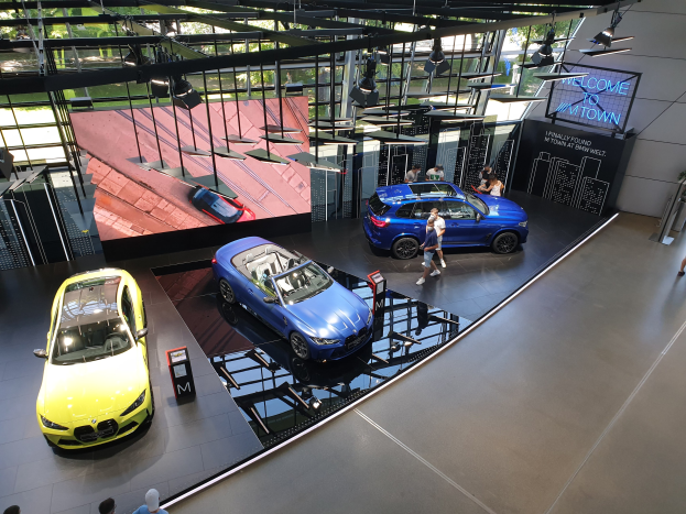 Eine Gruppe von Menschen, die im BMW Museum in München stehen, mit Autos zur Linken und einem Schild mit der Aufschrift "BMW Museum München" zur Rechten, mit Blick auf eine Außenansicht von Büschen, Gebüuden und Himmel durch Glasfenster.