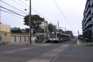 Straßenbahn fährt durch eine Straße mit hohen Gebäuden, Strommasten, Fußgängern, Bäumen und einem klaren blauen Himmel.