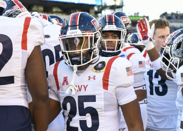 Gruppe von Männern in Fußballuniformen auf einem Feld mit Helmen und Handschuhen, mit einer unscharfen Menge, einer Anzeigetafel mit der Aufschrift "Auburn vs Virginia Tech - 11/28/15 College Football Pick, Odds, and Prediction" und Himmel im Hintergrund.