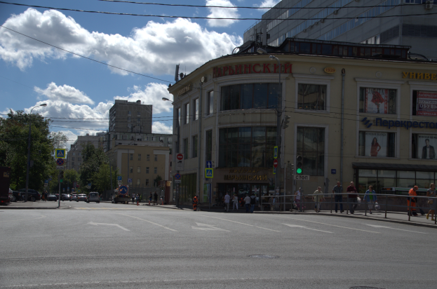 Eine Stadtstraße mit Fußgängern, hohe Gebäude mit Fenstern, Schilder, Ampeln, ein Metallzaun, Straßenlaternen, Fahrzeuge, Bäume und ein bewölkter Himmel.