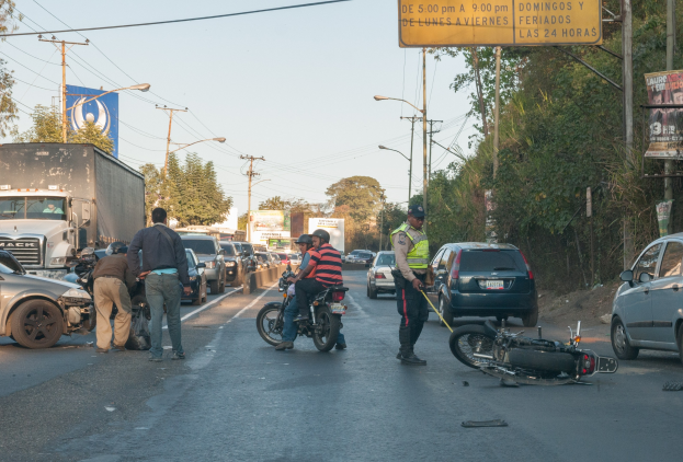 Gruppe von Menschen um ein verunglücktes Motorrad am Stra├čenrand mit mehreren Fahrzeugen, darunter ein Lastwagen, im Hintergrund und umgeben von Infrastruktur.