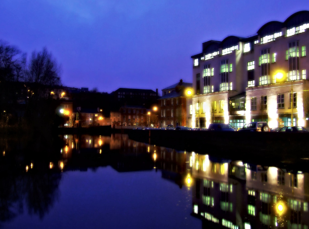 Ein Körper von Wasser im Vordergrund mit Bäumen, Gebäuden, Straßenlaternen und einem Himmel im Hintergrund, mit Autos auf einer Straße sichtbar.