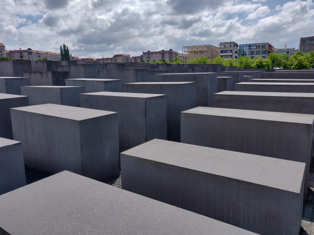 Ein Feld aus Betonbl√∂cken h√∂chsten variierenden, die in einem Gittermuster angeordnet sind, mit B√§umen und Geb√§uden im Hintergrund und einem bew√∂lktem Himmel √∂ben, das Holocaust-Mahnmal f√ľr die ermordeten Juden Europas in Berlin, Deutschland darstellend.