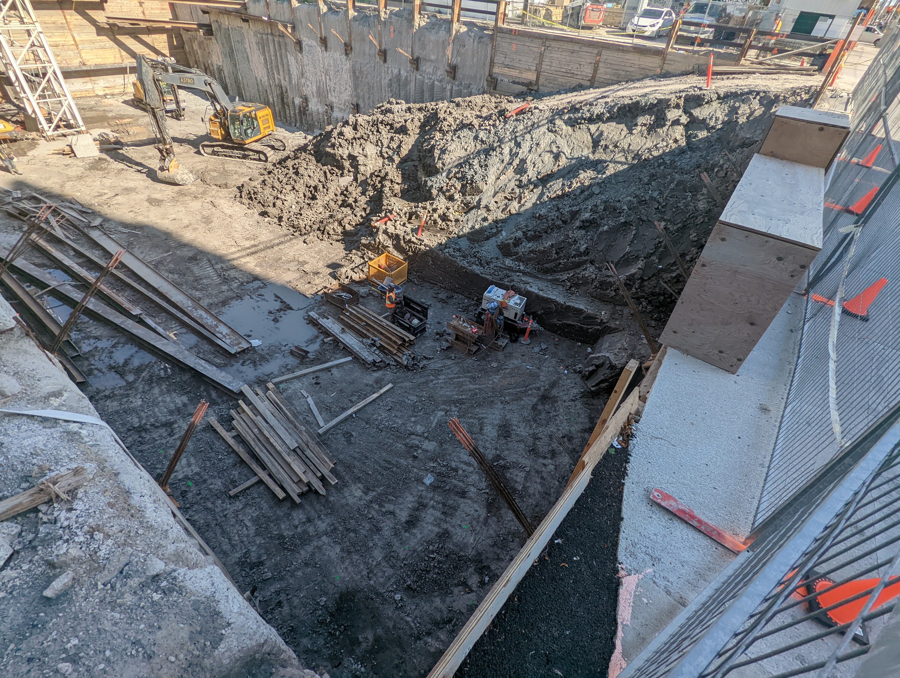 Baustelle mit einem großen Haufen Erde, umgeben von Kränen, Holzstöcken und anderem Equipment, abgegrenzt durch einen Maschendrahtzaun, mit Gebäuden, Fahrzeugen, Pfählen, Bäumen und Himmel im Hintergrund.