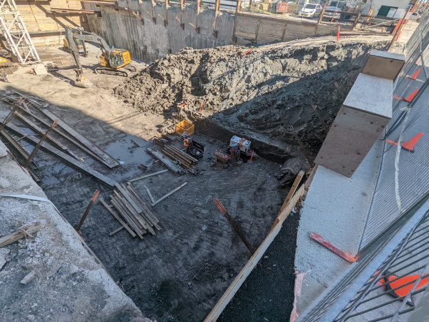 Baustelle mit einem großen Haufen Erde, umgeben von Kränen, Holzstöcken und anderem Equipment, abgegrenzt durch einen Maschendrahtzaun, mit Gebäuden, Fahrzeugen, Pfählen, Bäumen und Himmel im Hintergrund.