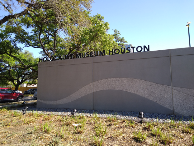 Das Holocaust-Museum in Houston, Texas, mit einer textbedeckten Wand, Steinen, Gras, Pflanzen, einer Baumgruppe, Fahrzeugen auf der Straße und einem Laternenpfahl.