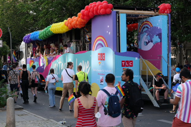 Eine Gruppe von Menschen, die eine Straße entlanggehen, neben einem Lastwagen mit bunten Ballons, mit Schildern an Pfosten an der Straße und Bäumen und Gebäuden im Hintergrund, was auf eine Pride-Parade in Paris hindeutet.