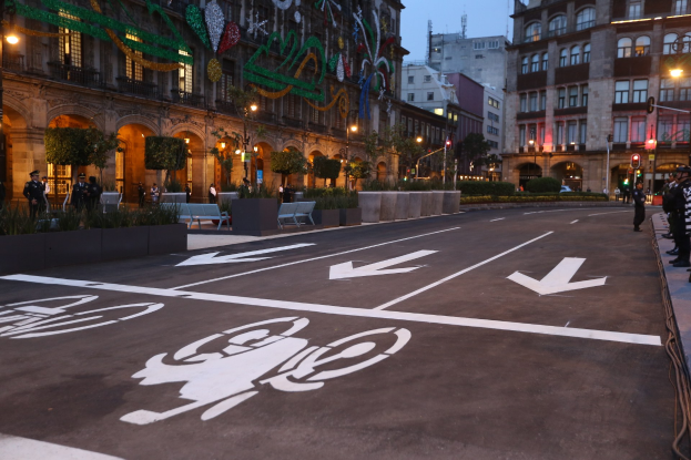 Stadtstraße mit einem Fahrradweg, umgeben von Gebäuden, Straßenmöbeln, Fußgängern, Grünflächen und einem klaren blauen Himmel.