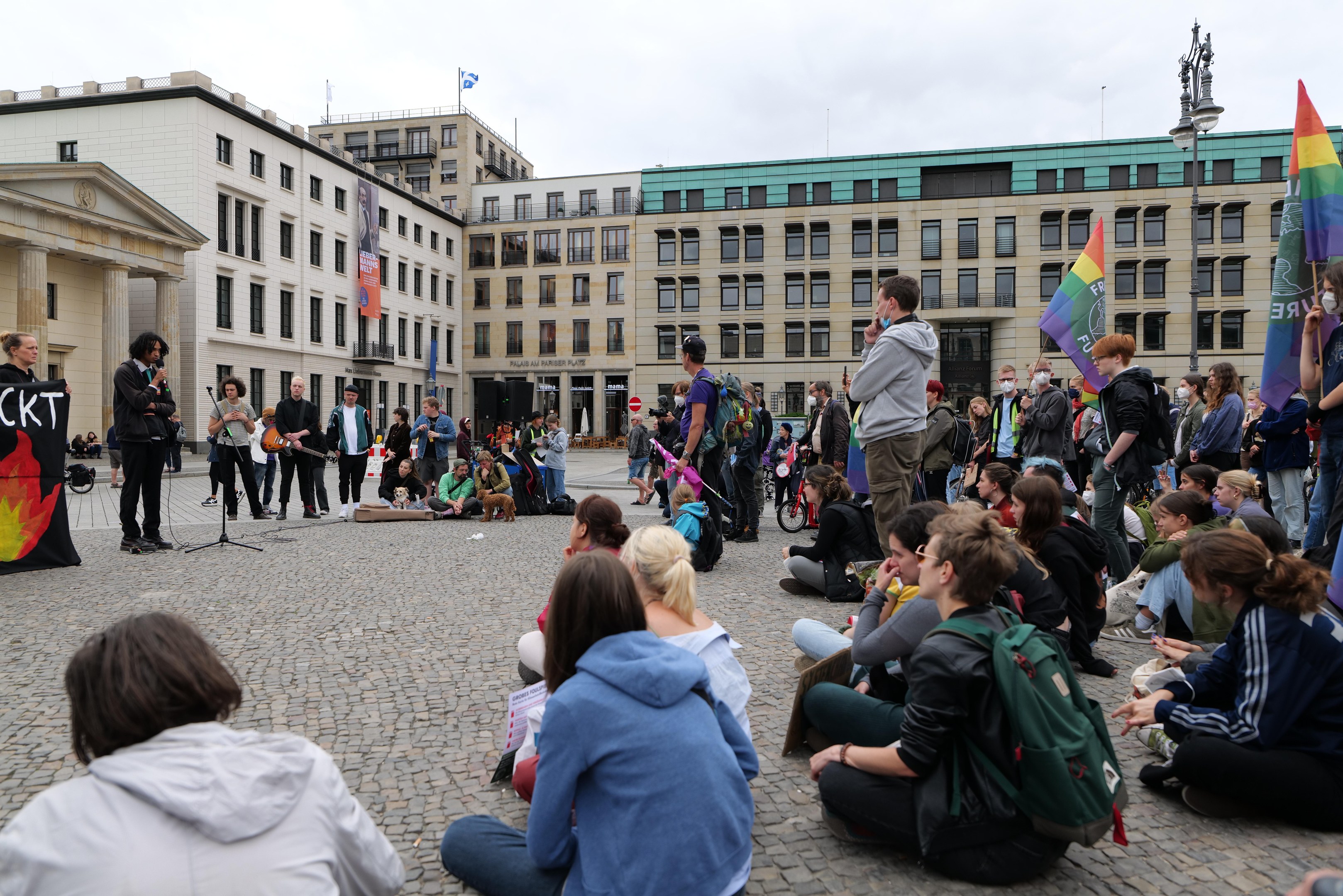 Eine Gruppe von Menschen, die auf dem Boden vor einer Menge sitzen, die Flaggen und Plakate hölt, während einer Anti-Schwulen-Demo in Berlin. Im Hintergrund sind eine Statue, ein Mikrofon und Gebäude zu sehen.