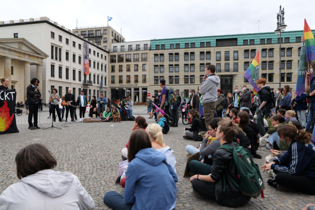 Eine Gruppe von Menschen, die auf dem Boden vor einer Menge sitzen, die Flaggen und Plakate hölt, während einer Anti-Schwulen-Demo in Berlin. Im Hintergrund sind eine Statue, ein Mikrofon und Gebäude zu sehen.