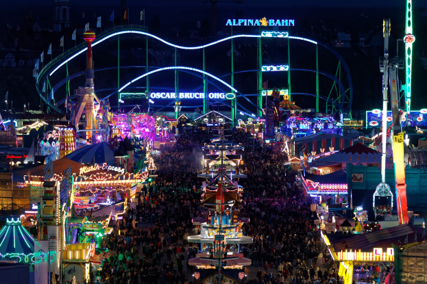 Eine große Menschenmenge steht um einen hell erleuchteten Karneval in der Nacht herum, umgeben von Vergnügungsgeräten, Pfählen, Fahnen und Namensschildern, mit einem dunklen Hintergrund, der eine festliche Atmosphäre schafft.