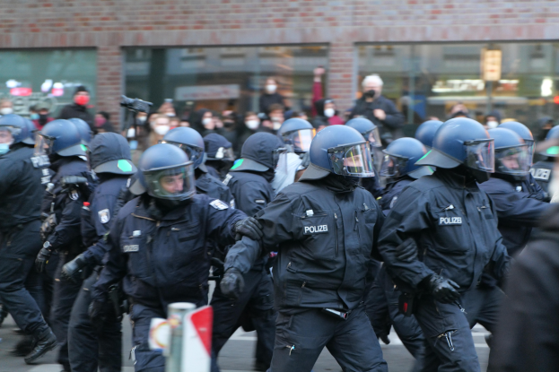 Eine Gruppe von Polizisten in Einsatzausrüstung, darunter Helme und Handschuhe, geht eine Straße entlang mit einem unscharfen Hintergrund, der ein Gebäude mit Glaswänden und eine Schilderstange auf einem Pfahl zeigt.