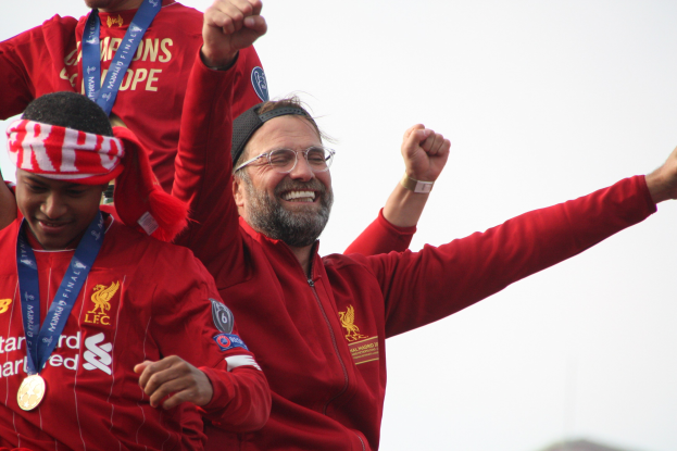 Liverpool-Trainer Jürgen Klopp feiert mit Spielern und Staff nach dem Gewinn der UEFA Champions League, trägt eine rote Jacke mit zwei Medaillienträgern und einem sichtbaren Himmel im Hintergrund.