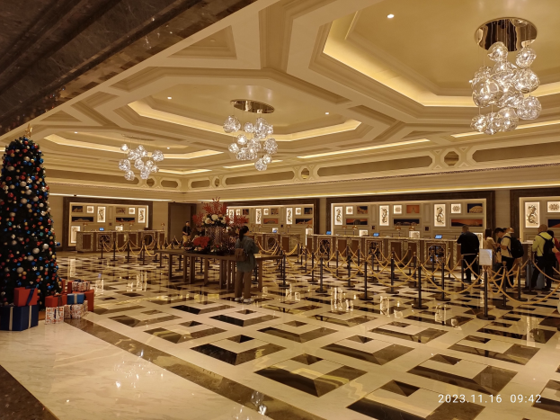 Ein geräumiger Eingangsbereich mit einem zentralen Weihnachtsbaum, umgeben von Geschenkboxen und festlichen Dekorationen, mit Menschen, Absperrpollern mit Seilen, Blumenbouquets auf Tischen, gerahmten Fotos an den Wänden und beleuchteten Deckenlampen und Kronleuchtern.