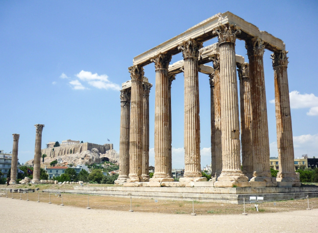 Alter Tempel von Olympie in Athen, Griechenland, mit hohen Säulen, umgeben von Bäumen und Felsen, und eine Burg sichtbar im bewölkten Himmel.