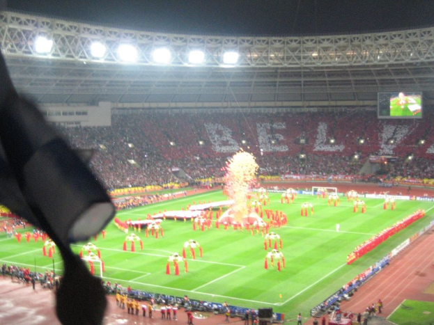 Ein Stadion mit Flutlicht, Eisengittern, Zuschauern, stehenden Personen, Werbetafeln, Anzeigetafeln, Sicherheitskameras und dekorativen Flaggen.