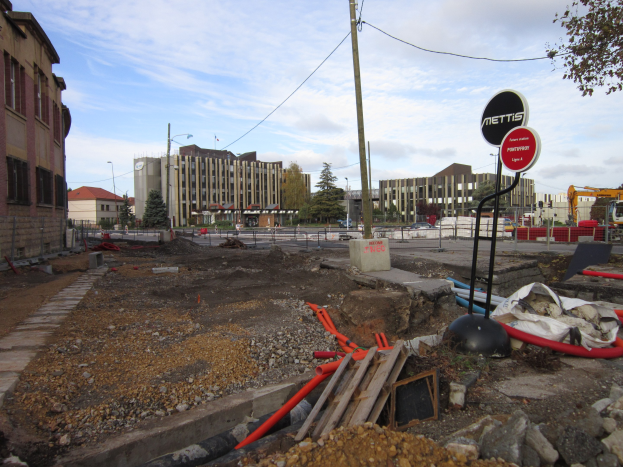Baustelle mit verstreuten Materialien wie Steinen und Rohren, Hinweisschilder an Pfählen, ein Zaun, Fahrzeuge, Bäume, Gebäude mit Fenstern, Laternenpfähle und ein bewölkter Himmel im Hintergrund.
