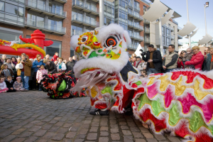 Ein lebendiges chinesisches Neujahrsfest in Amsterdam mit einer Löwen-Tanz-Performance vor einer Zuschauermenge, darunter einige, die das Ereignis fotografieren, vor einer Kulisse aus Gebäuden, Laternenmasten und einem klaren blauen Himmel.