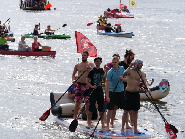 Eine Gruppe von Menschen auf Paddle-Boards im Wasser, einige halten Paddel, mit Booten und Fahnen im Hintergrund auf der 2016 Stand Up Paddleboard Festival.