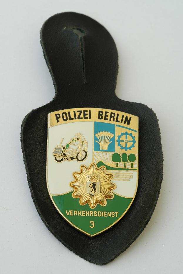 Ein schildförmiges Polizeibadgebnis mit "Polizei Berlin" in schwarzer fettgeschriebener Schrift, das ein weißes Stern auf einem blauen Hintergrund und einen weißen Rand hat, das auf einer Oberfläche liegt.