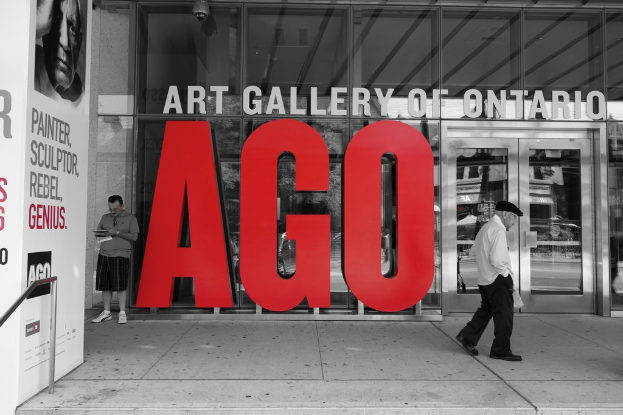 Ein Mann steht mit einem Notizbuch in der Nähe der Glaswand der Kunstgalerie von Ontario und schreibt darin, während ein anderer Mann in der Nähe geht.