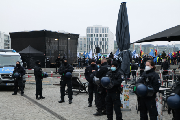 Polizeibeamte in Uniform stehen hinter Absperrungen und blicken auf eine Menge von Demonstranten, die Fahnen schwenken und Helme und Masken tragen.