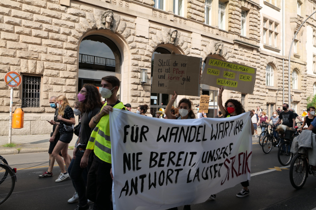 Eine Gruppe von Menschen marschiert bei einer Protestaktion gegen die Friedensbewegung in Berlin, einige halten Schilder und andere fahren Fahrräder, im Hintergrund ein Gebäude mit Fenstern, Bögen, Säulen und Skulpturen.