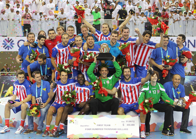 Eine Gruppe von Menschen in Sportkleidung, einige halten eine Trophäe und Blumen, stehen vor einem Publikum mit Geländern und Schildern im Hintergrund.