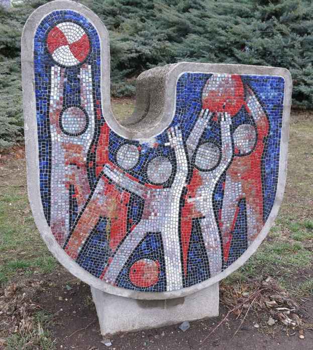 Eine Mosaikbank in einem Park, umgeben von Bäumen unter einem klaren blauen Himmel, mit farbigen Fliesen, die ein einzigartiges Design schaffen.