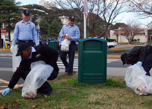 Drei Polizeibeamte in Mützen und Handschuhen sammeln Müll an einer Straßenböschung mit Bäumen, Gebäuden, Fahrzeugen und einem Mülleimer im Hintergrund.