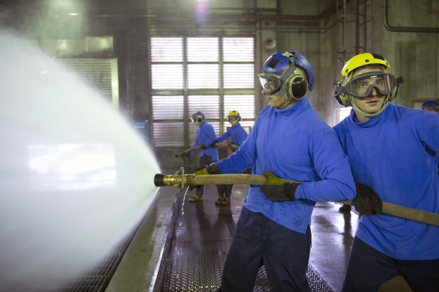 Eine Gruppe von Männern in blauen Hemden und gelben Helmen arbeitet an Maschinen, wobei einer Wasser auf den Boden sprüht, in einer Fabrik mit sichtbaren Rohren, Fenstern und Lampen.