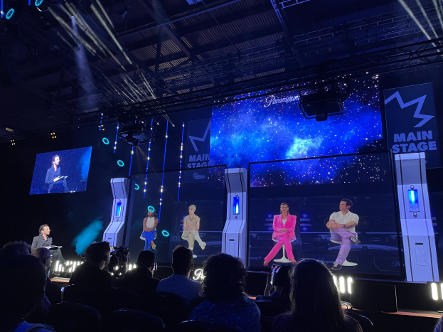 Eine Gruppe von Menschen auf einer hell erleuchteten Bühne mit Bildschirmen, die einen Redner mit Mikrofon und ein Publikum vorne zeigt, unter einer Decke mit Beleuchtungsequipment.