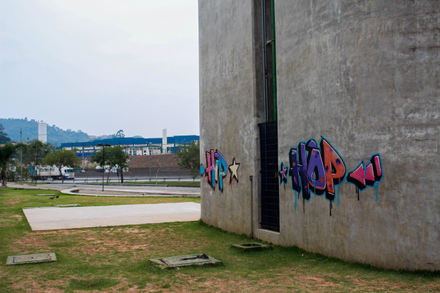 Betongebäude mit Graffiti, umgeben von Gras, Bäumen, Masten, Fahrzeugen, anderen Gebäuden, Hügeln und einem klaren blauen Himmel.
