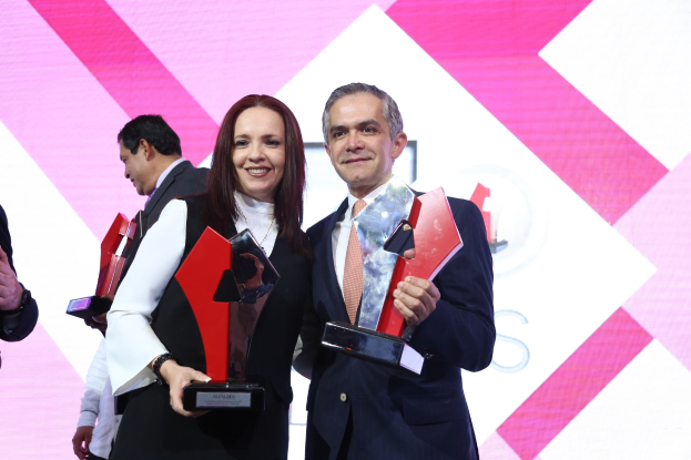 Zwei strahlende Menschen mit Trophäen auf einer Bühne bei einer Preisverleihung, mit einem Bildschirm im Hintergrund.