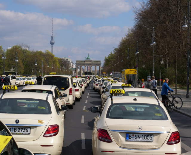 Eine lange Reihe von Taxis, die entlang einer belebten Straße in Berlin, Deutschland, geparkt sind, mit Fahrzeugen, Radfahrern und Fußgängern in der Nähe, flankiert von Bäumen und Laternenmasten und Gebäuden, einem Bogen und einem Turm im Hintergrund unter einem bewölkten Himmel.