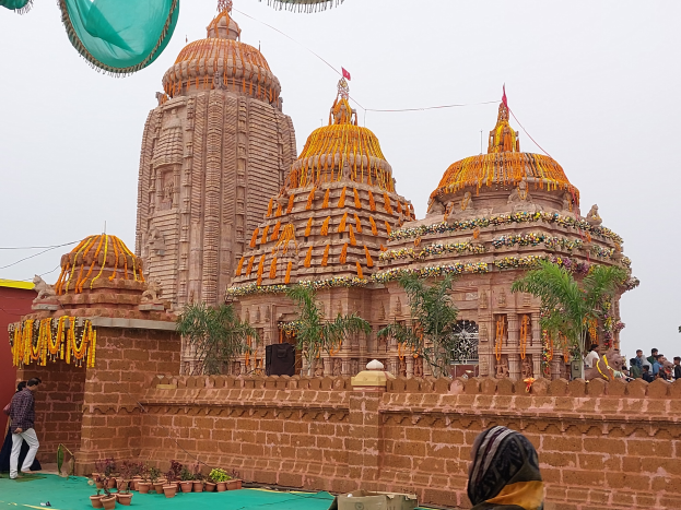 Eine Gruppe von Menschen vor einem Hindu-Tempel mit bunten Girlanden und Flaggen, umgeben von Grün unter einem hellblauen Himmel, die sich um einen grünen Teppich versammelt haben, während sie das Jagannath-Fest feiern.