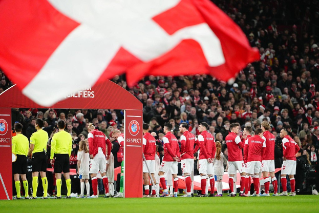 Eine Gruppe von Menschen auf einem Fußballfeld mit einer roten und weißen Fahne im Vordergrund, einem Bogen mit dem Text "Bayern München vs. Bayern München Wetten & Vorschau" im Hintergrund und einer großen Menge im Stadion.