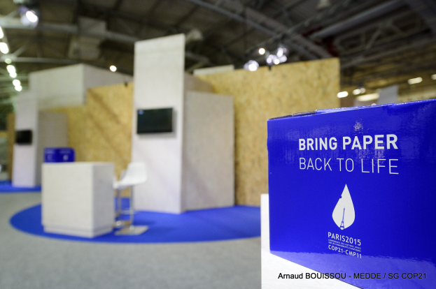 Ein Messestand mit einem blauen und weißen "Bring Paper Back to Life"-Schild auf der rechten Seite, mit unscharfen Stühlen, Tischen, einem wandmontierten Fernseher und Deckenleuchten im Hintergrund.