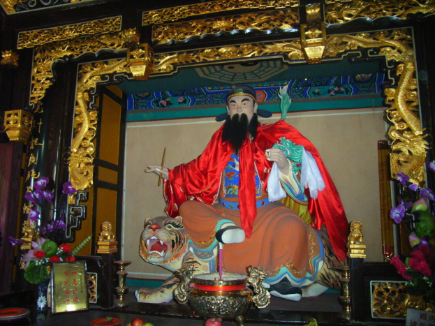 Eine Statue eines chinesischen Mönchs in buntem Gewand sitzt auf einem Tiger innerhalb eines Bogens mit goldenen Verzierungen, vorne ein Blumenkübel und in der Mitte eine Schale.