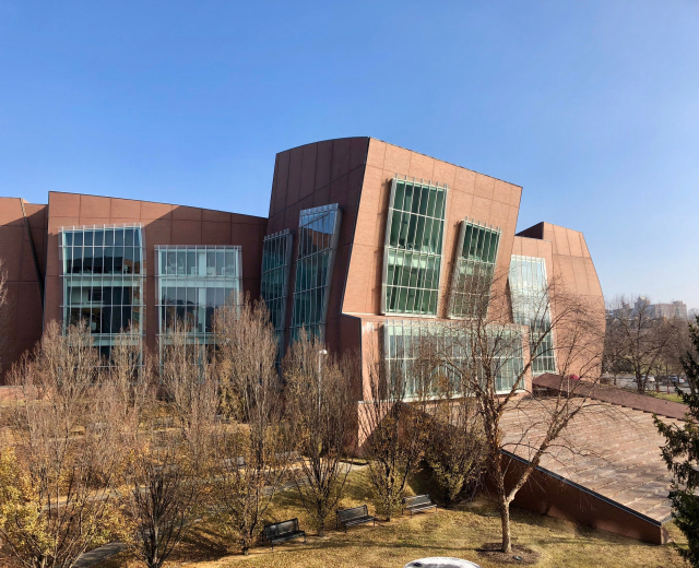 Großes Gebäude mit vielen Fenstern, umgeben von Bäumen, Bänken, Gras und einem Laternenmast, mit anderen Gebäuden und einem klaren blauen Himmel im Hintergrund; als der neue Campus der University of Illinois at Chicago bezeichnet.