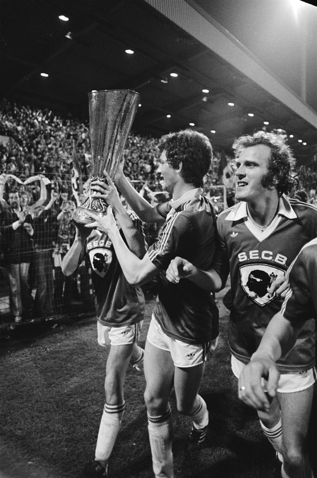 Eine Gruppe von Männern in Schwarz-Weiß-Kleidung hält einen Pokal auf einem Fußballfeld hoch und feiert ihren Sieg im FA-Cup-Finale, umgeben von einer Menge hinter einem Zaun unter Stadionbeleuchtung.